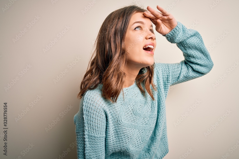 Young blonde girl wearing casual blue winter sweater over isolated background very happy and smiling looking far away with hand over head. Searching concept. Young blonde girl wearing casual blue winter sweater over isolated background very happy and smiling looking far away with hand over head. Searching concept.