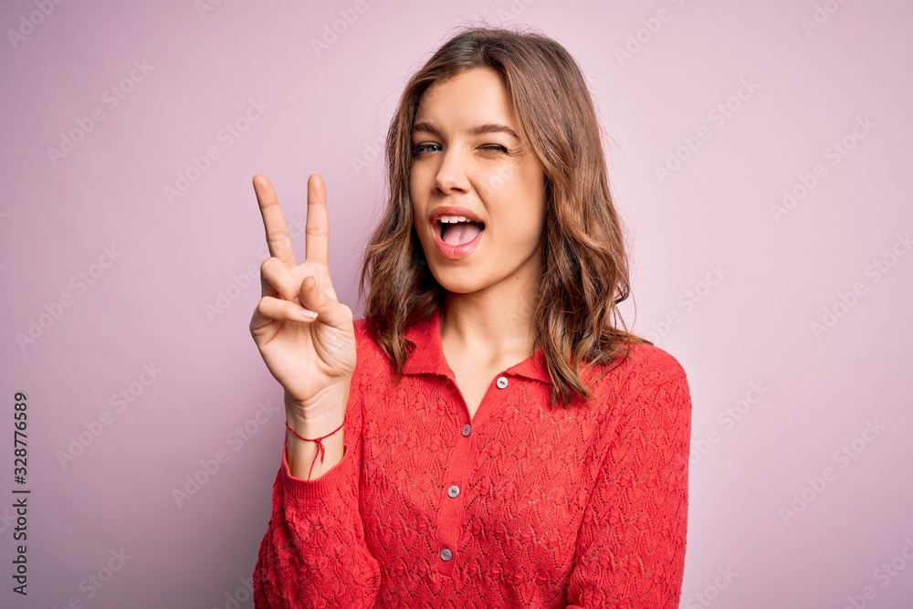 Young beautiful blonde casual girl over pink isolated background smiling with happy face winking at the camera doing victory sign. Number two. Young beautiful blonde casual girl over pink isolated background smiling with happy face winking at the camera doing victory sign. Number two.