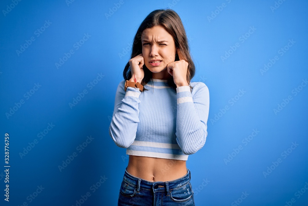 Young beautiful blonde girl wearing casual sweater standing over blue isolated background covering ears with fingers with annoyed expression for the noise of loud music. Deaf concept. Young beautiful blonde girl wearing casual sweater standing over blue isolated background covering ears with fingers with annoyed expression for the noise of loud music. Deaf concept.
