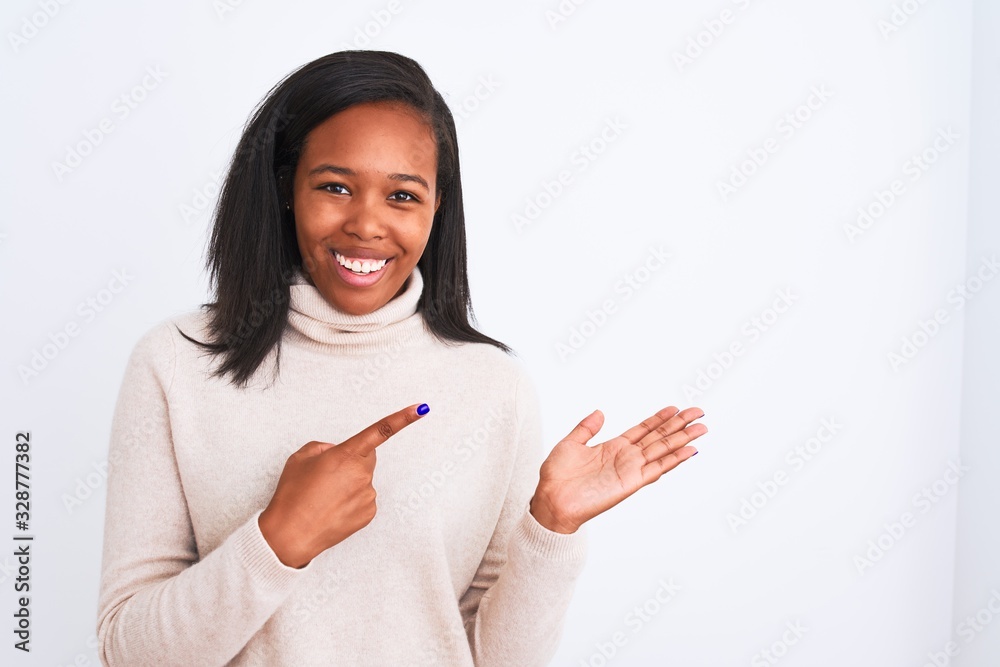 Beautiful young african american woman wearing turtleneck sweater over isolated background amazed and smiling to the camera while presenting with hand and pointing with finger. Beautiful young african american woman wearing turtleneck sweater over isolated background amazed and smiling to the camera while presenting with hand and pointing with finger.