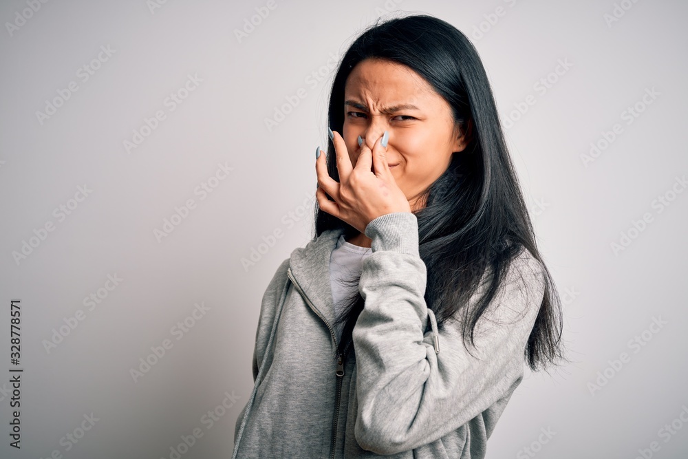 Young beautiful chinese sporty woman wearing sweatshirt over isolated white background smelling something stinky and disgusting, intolerable smell, holding breath with fingers on nose. Bad smell Young beautiful chinese sporty woman wearing sweatshirt over isolated white background smelling something stinky and disgusting, intolerable smell, holding breath with fingers on nose. Bad smell
