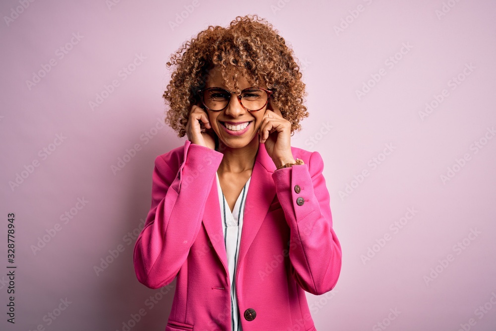 Young african american businesswoman wearing glasses standing over pink background covering ears with fingers with annoyed expression for the noise of loud music. Deaf concept. Young african american businesswoman wearing glasses standing over pink background covering ears with fingers with annoyed expression for the noise of loud music. Deaf concept.
