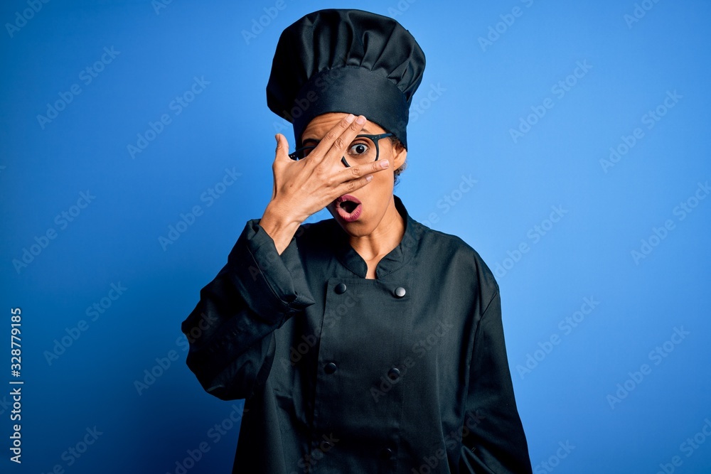 Young african american chef woman wearing cooker uniform and hat over blue background peeking in shock covering face and eyes with hand, looking through fingers with embarrassed expression. Young african american chef woman wearing cooker uniform and hat over blue background peeking in shock covering face and eyes with hand, looking through fingers with embarrassed expression.