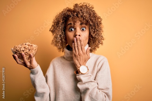Young african american woman with curly hair holding bowl with healthy peanuts cover mouth with hand shocked with shame for mistake, expression of fear, scared in silence, secret concept
