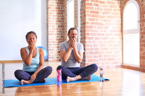 Middle age sporty couple sitting on mat doing stretching yoga exercise at gym bored yawning tired covering mouth with hand. Restless and sleepiness.