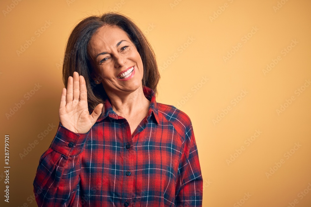 Middle age beautiful woman wearing casual shirt standing over isolated yellow background Waiving saying hello happy and smiling, friendly welcome gesture Middle age beautiful woman wearing casual shirt standing over isolated yellow background Waiving saying hello happy and smiling, friendly welcome gesture