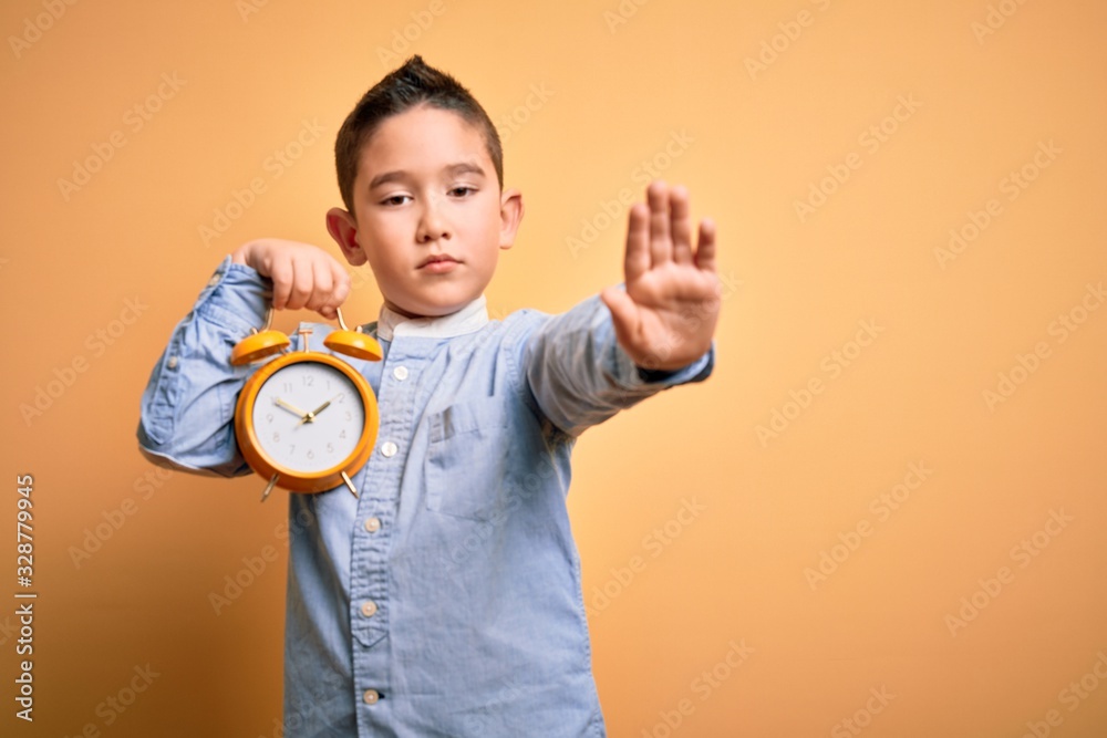 Young little boy kid holding classic bell alarm clock over isolated yellow background with open hand doing stop sign with serious and confident expression, defense gesture Young little boy kid holding classic bell alarm clock over isolated yellow background with open hand doing stop sign with serious and confident expression, defense gesture