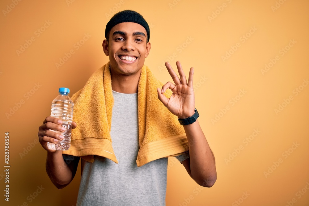 Young handsome african american sportsman wearing towel drinking bottle of water doing ok sign with fingers, excellent symbol Young handsome african american sportsman wearing towel drinking bottle of water doing ok sign with fingers, excellent symbol