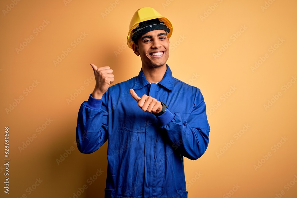 Young handsome african american worker man wearing blue uniform and security helmet Pointing to the back behind with hand and thumbs up, smiling confident