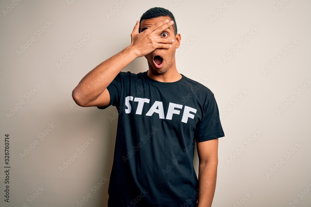 Young handsome african american worker man wearing staff uniform over white background peeking in shock covering face and eyes with hand, looking through fingers with embarrassed expression. Young handsome african american worker man wearing staff uniform over white background peeking in shock covering face and eyes with hand, looking through fingers with embarrassed expression.