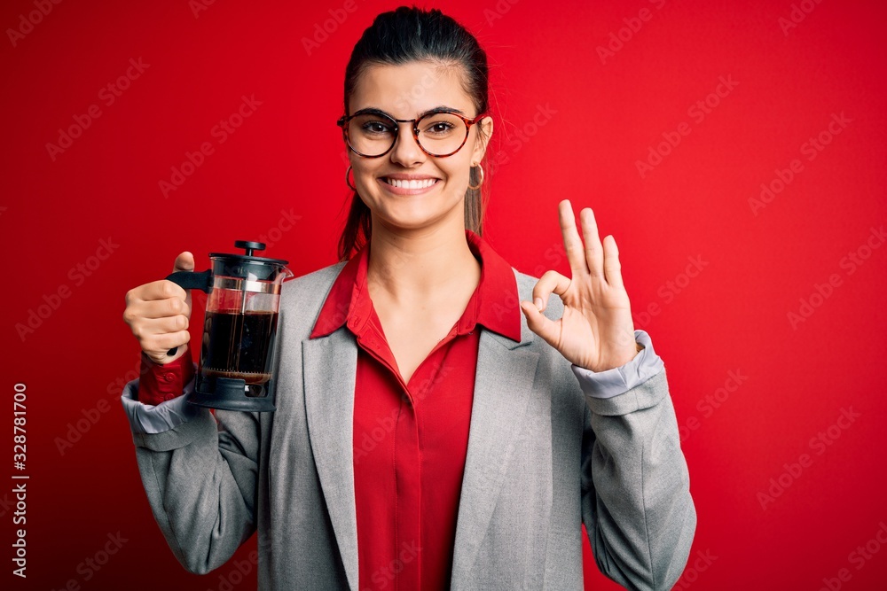Young beautiful brunette woman doing coffe holding french coffeemaker over red background doing ok sign with fingers, excellent symbol Young beautiful brunette woman doing coffe holding french coffeemaker over red background doing ok sign with fingers, excellent symbol
