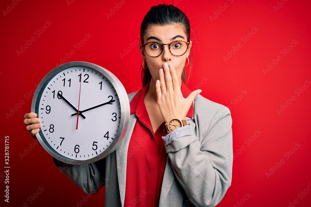 Young beautiful brunette businesswoman doing countdown holding big clock cover mouth with hand shocked with shame for mistake, expression of fear, scared in silence, secret concept Young beautiful brunette businesswoman doing countdown holding big clock cover mouth with hand shocked with shame for mistake, expression of fear, scared in silence, secret concept