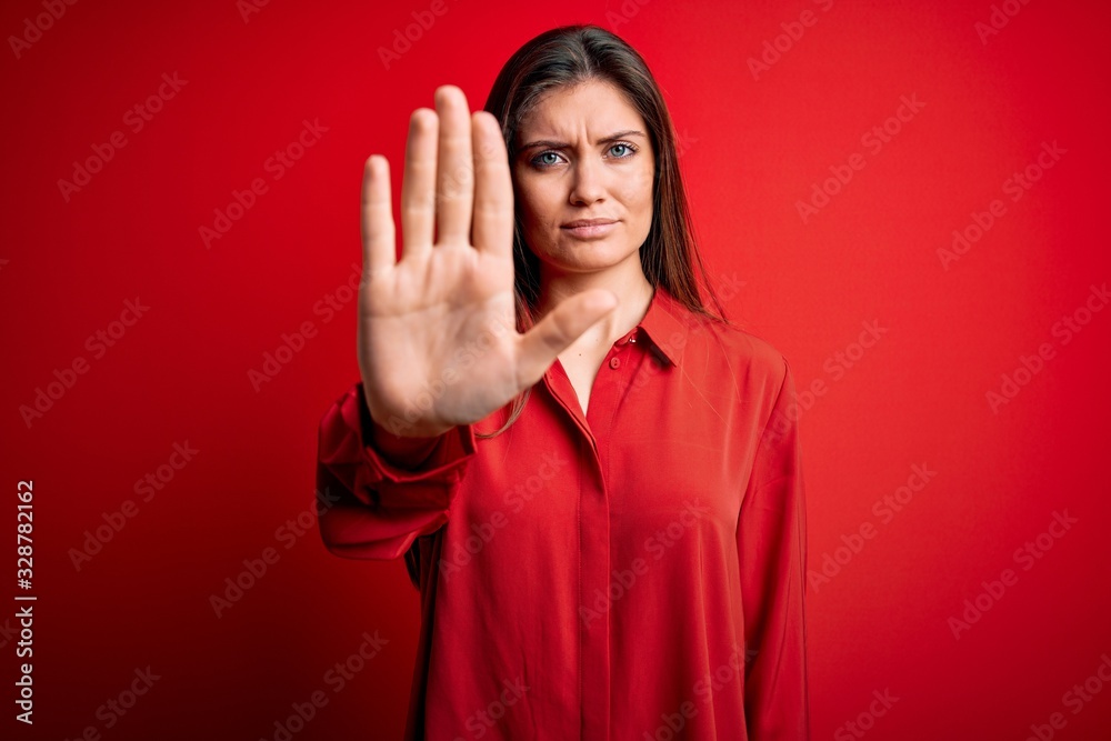Young beautiful woman with blue eyes wearing casual shirt standing over red background doing stop sing with palm of the hand. Warning expression with negative and serious gesture on the face.
