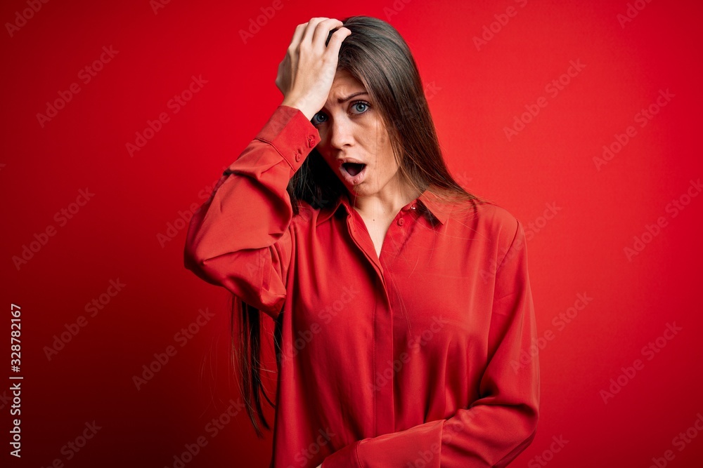 Young beautiful woman with blue eyes wearing casual shirt standing over red background surprised with hand on head for mistake, remember error. Forgot, bad memory concept. Young beautiful woman with blue eyes wearing casual shirt standing over red background surprised with hand on head for mistake, remember error. Forgot, bad memory concept.