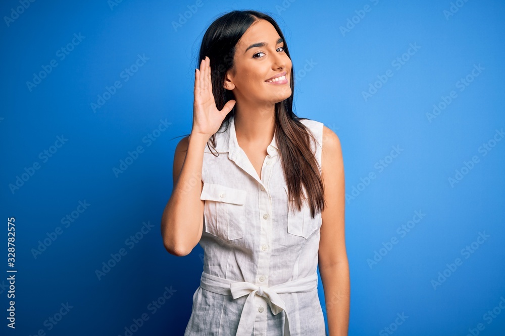 Young beautiful brunette woman wearing casual dress over isolated blue background smiling with hand over ear listening an hearing to rumor or gossip. Deafness concept. Young beautiful brunette woman wearing casual dress over isolated blue background smiling with hand over ear listening an hearing to rumor or gossip. Deafness concept.
