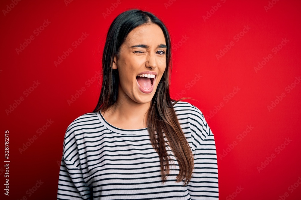 Young beautiful brunette woman wearing casual striped t-shirt over red background winking looking at the camera with sexy expression, cheerful and happy face. Young beautiful brunette woman wearing casual striped t-shirt over red background winking looking at the camera with sexy expression, cheerful and happy face.