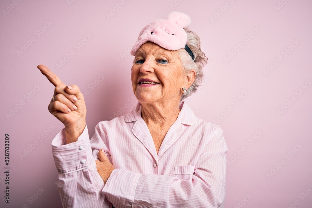 Senior beautiful woman wearing sleep mask and pajama over isolated pink background with a big smile on face, pointing with hand and finger to the side looking at the camera. Senior beautiful woman wearing sleep mask and pajama over isolated pink background with a big smile on face, pointing with hand and finger to the side looking at the camera.