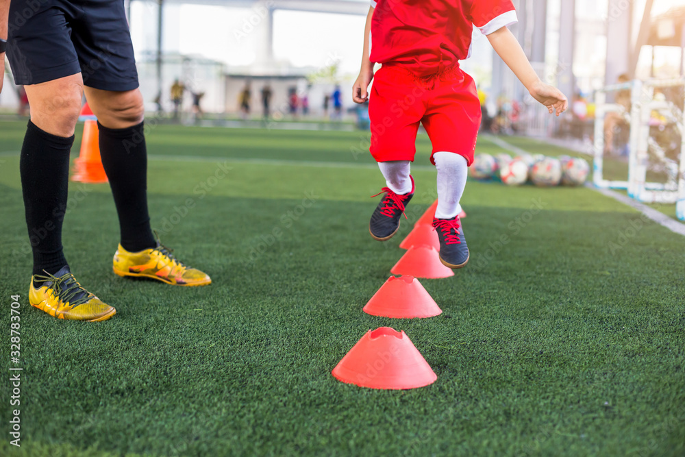 Obraz premium Kid soccer jogging and jump between cone marker with coach standing to coaching in soccer academy. Coach is coaching kid soccer player to jogging and jump between ladder drills for exercises.