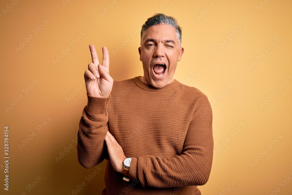 Middle age handsome grey-haired man wearing casual sweater over yellow background smiling with happy face winking at the camera doing victory sign. Number two. - obrazy, fototapety, plakaty 