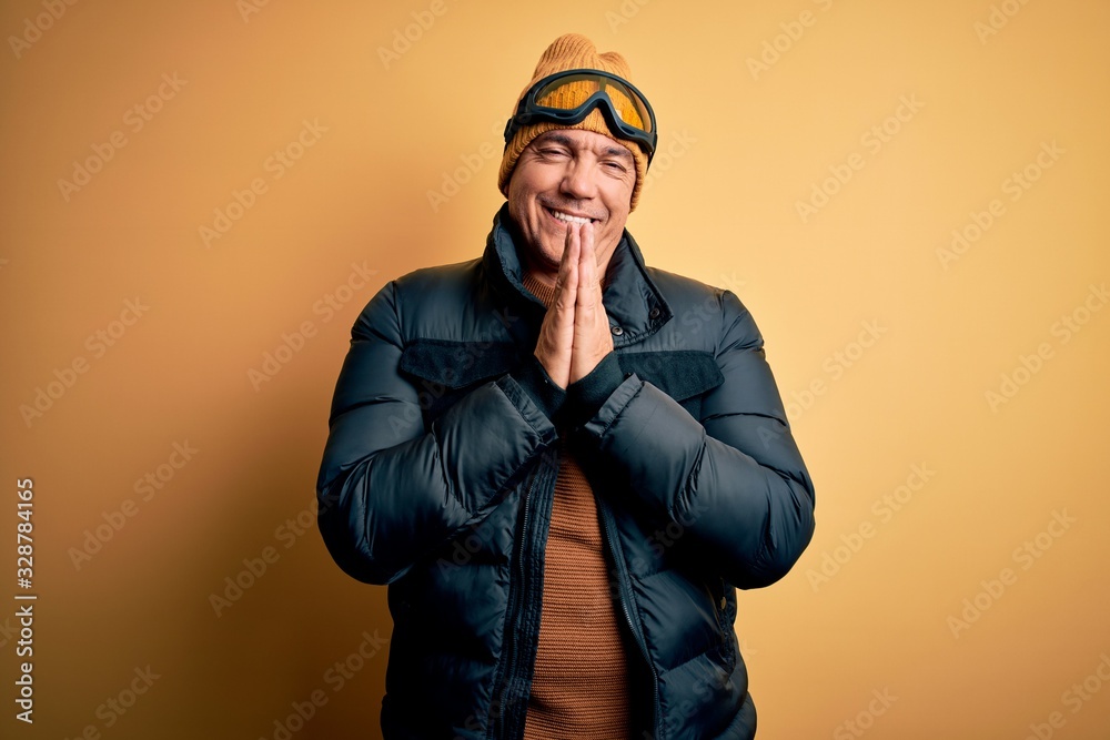 Middle age handsome grey-haired skier man on vacation wearing ski goggles praying with hands together asking for forgiveness smiling confident.