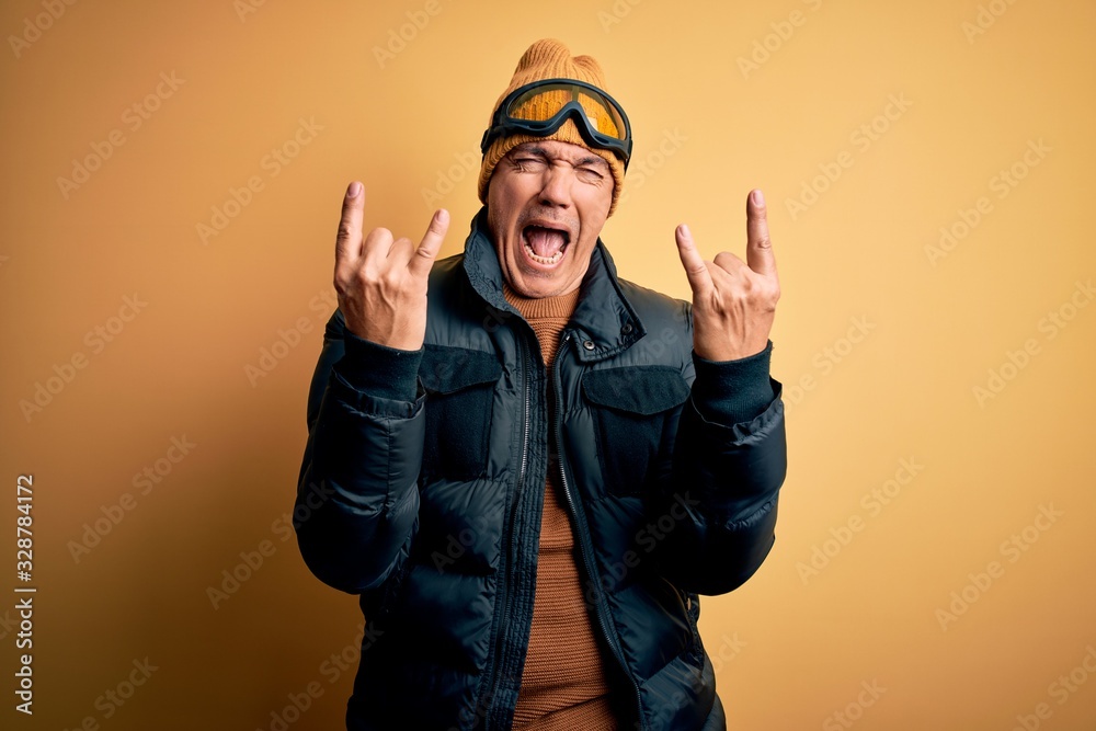 Middle age handsome grey-haired skier man on vacation wearing ski goggles shouting with crazy expression doing rock symbol with hands up. Music star. Heavy concept. Middle age handsome grey-haired skier man on vacation wearing ski goggles shouting with crazy expression doing rock symbol with hands up. Music star. Heavy concept.