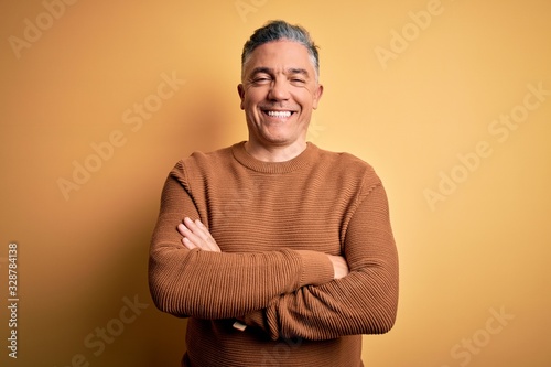 Middle age handsome grey-haired man wearing casual sweater over yellow background happy face smiling with crossed arms looking at the camera. Positive person.
