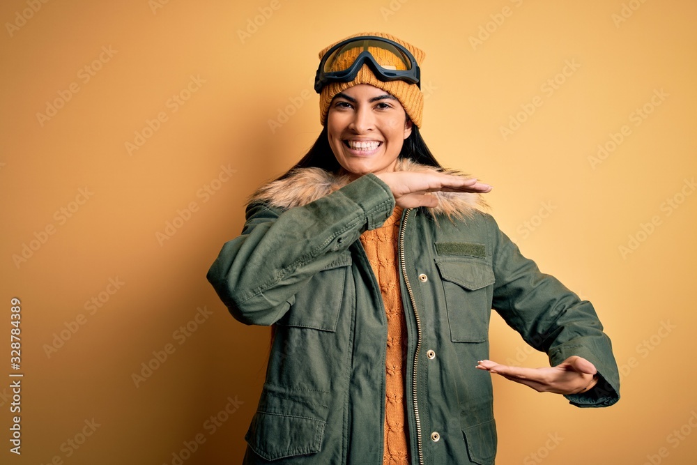 Young beautiful hispanic woman wearing ski glasses and coat for winter weather gesturing with hands showing big and large size sign, measure symbol. Smiling looking at the camera. Measuring concept. Young beautiful hispanic woman wearing ski glasses and coat for winter weather gesturing with hands showing big and large size sign, measure symbol. Smiling looking at the camera. Measuring concept.
