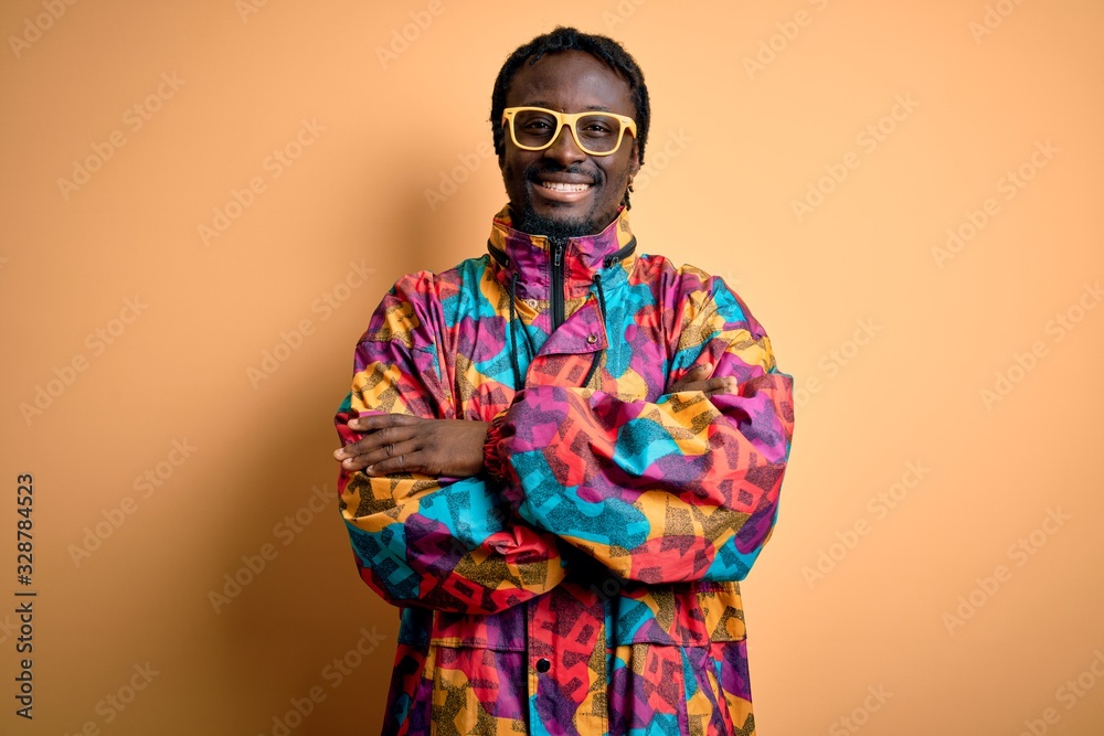 Handsome african american man wearing colorful coat and glasses over yellow background happy face smiling with crossed arms looking at the camera. Positive person.
