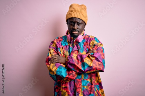 Young handsome african american man wearing colorful coat and cap over pink background skeptic and nervous, disapproving expression on face with crossed arms. Negative person.