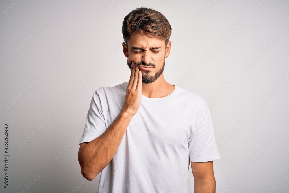 Young handsome man with beard wearing casual t-shirt standing over white background touching mouth with hand with painful expression because of toothache or dental illness on teeth. Dentist Young handsome man with beard wearing casual t-shirt standing over white background touching mouth with hand with painful expression because of toothache or dental illness on teeth. Dentist