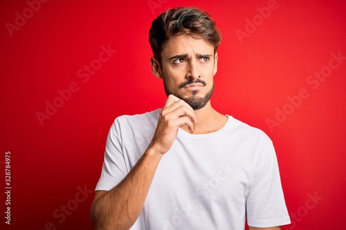Young handsome man with beard wearing casual t-shirt standing over red background with hand on chin thinking about question, pensive expression. Smiling with thoughtful face. Doubt concept.
