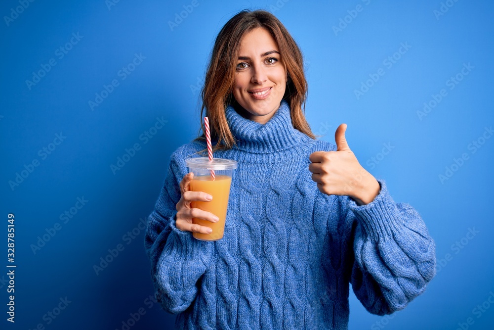 Young beautiful brunette woman drinking healthy orange juice over blue background happy with big smile doing ok sign, thumb up with fingers, excellent sign Young beautiful brunette woman drinking healthy orange juice over blue background happy with big smile doing ok sign, thumb up with fingers, excellent sign