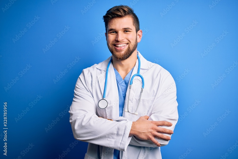 Young blond doctor man with beard and blue eyes wearing white coat and stethoscope happy face smiling with crossed arms looking at the camera. Positive person. Young blond doctor man with beard and blue eyes wearing white coat and stethoscope happy face smiling with crossed arms looking at the camera. Positive person.