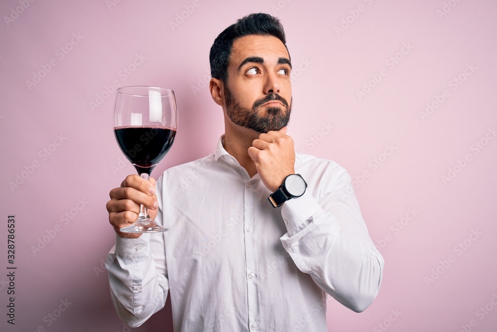 Young handsome man with beard drinking glass of red wine over isolated pink background serious face thinking about question, very confused idea Young handsome man with beard drinking glass of red wine over isolated pink background serious face thinking about question, very confused idea