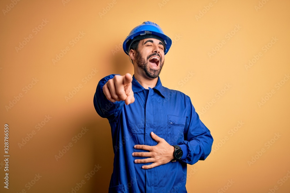 Mechanic man with beard wearing blue uniform and safety helmet over yellow background laughing at you, pointing finger to the camera with hand over body, shame expression