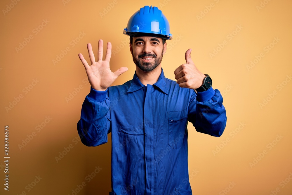 Mechanic man with beard wearing blue uniform and safety helmet over yellow background showing and pointing up with fingers number six while smiling confident and happy.