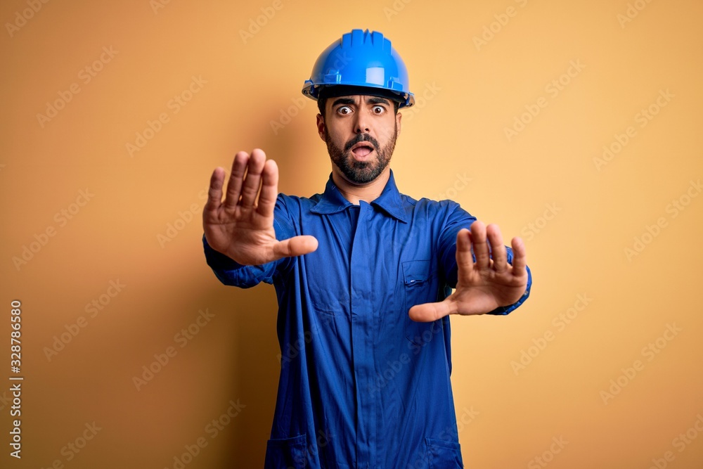 Mechanic man with beard wearing blue uniform and safety helmet over yellow background doing stop gesture with hands palms, angry and frustration expression