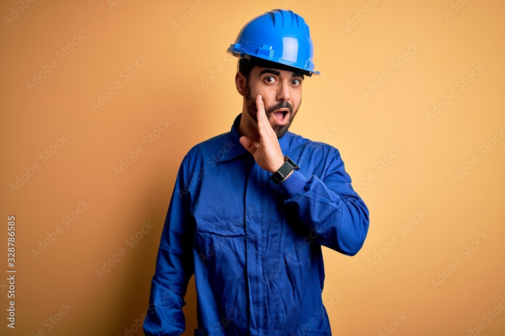 Mechanic man with beard wearing blue uniform and safety helmet over yellow background hand on mouth telling secret rumor, whispering malicious talk conversation