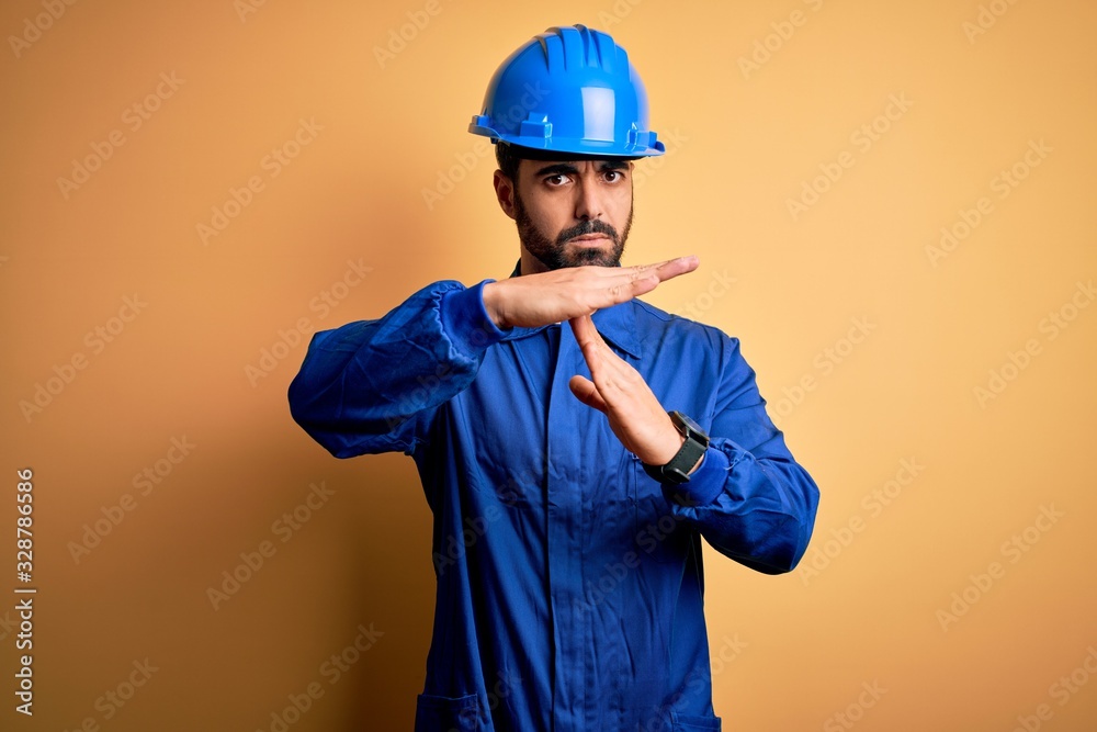 Mechanic man with beard wearing blue uniform and safety helmet over yellow background Doing time out gesture with hands, frustrated and serious face