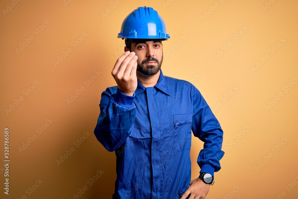 Mechanic man with beard wearing blue uniform and safety helmet over yellow background Doing Italian gesture with hand and fingers confident expression