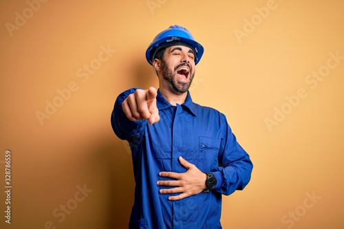 Mechanic man with beard wearing blue uniform and safety helmet over yellow background laughing at you  pointing finger to the camera with hand over body  shame expression