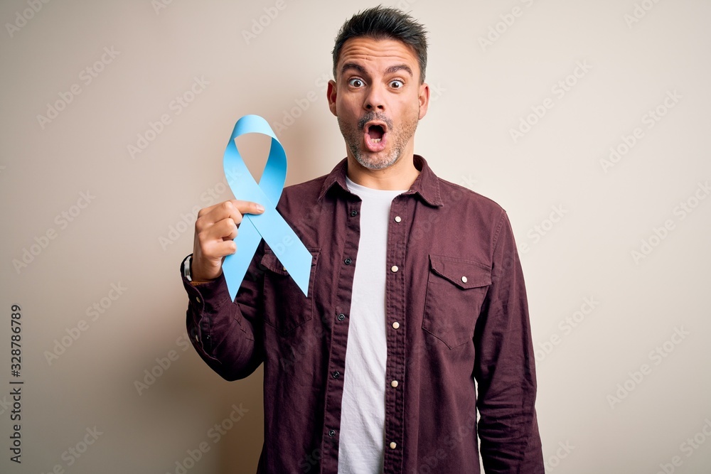 Young handsome man holding blue cancer ribbon over isolated white background scared in shock with a surprise face, afraid and excited with fear expression