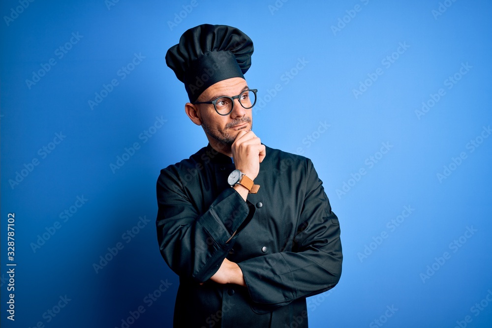 Young handsome chef man wearing cooker uniform and hat over isolated blue background with hand on chin thinking about question, pensive expression. Smiling with thoughtful face. Doubt concept. Young handsome chef man wearing cooker uniform and hat over isolated blue background with hand on chin thinking about question, pensive expression. Smiling with thoughtful face. Doubt concept.