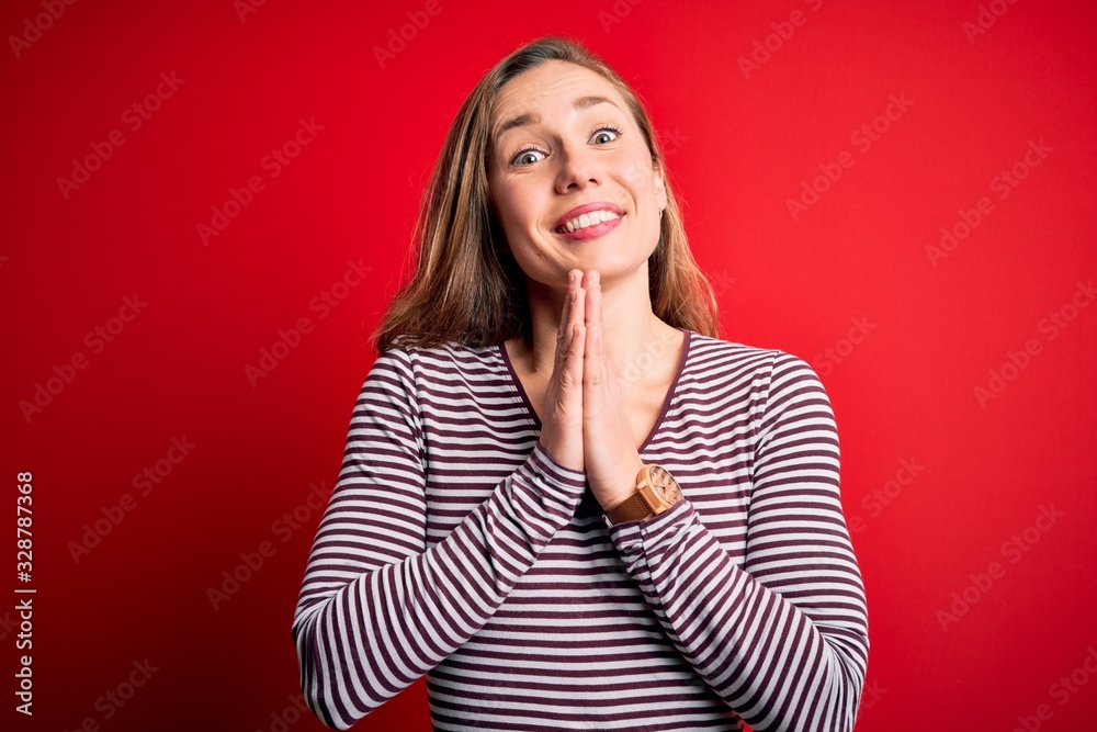 Young beautiful blonde woman wearing casual striped t-shirt over isolated red background praying with hands together asking for forgiveness smiling confident. Young beautiful blonde woman wearing casual striped t-shirt over isolated red background praying with hands together asking for forgiveness smiling confident.