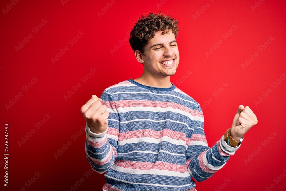 Young blond handsome man with curly hair wearing striped sweater over red background very happy and excited doing winner gesture with arms raised, smiling and screaming for success. Celebration Young blond handsome man with curly hair wearing striped sweater over red background very happy and excited doing winner gesture with arms raised, smiling and screaming for success. Celebration