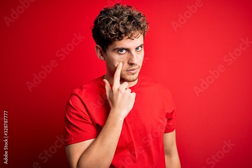 Young blond handsome man with curly hair wearing casual t-shirt over red background Pointing to the eye watching you gesture, suspicious expression