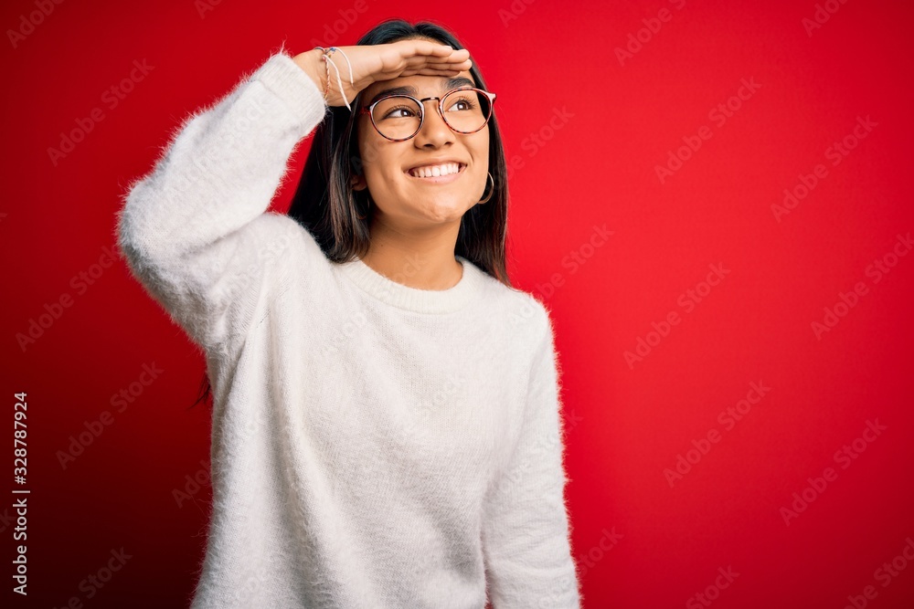 Young beautiful asian woman wearing casual sweater and glasses over red background very happy and smiling looking far away with hand over head. Searching concept. Young beautiful asian woman wearing casual sweater and glasses over red background very happy and smiling looking far away with hand over head. Searching concept.