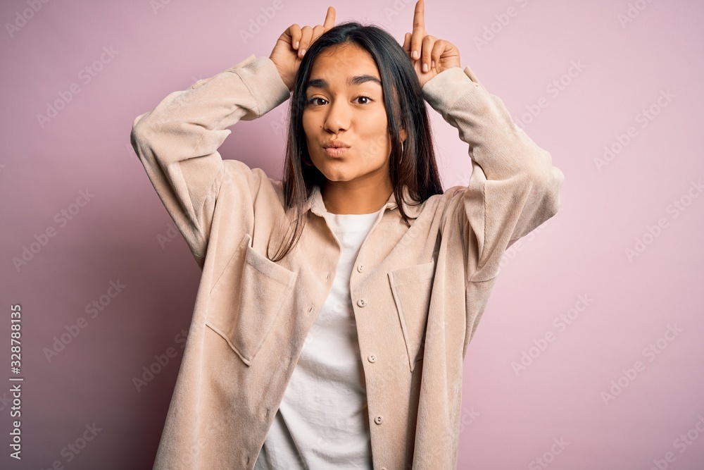 Young beautiful asian woman wearing casual shirt standing over pink background doing funny gesture with finger over head as bull horns Young beautiful asian woman wearing casual shirt standing over pink background doing funny gesture with finger over head as bull horns