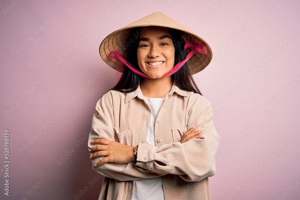 Young beautiful woman wearing traditional conical asian hat over isolated pink background happy face smiling with crossed arms looking at the camera. Positive person. Young beautiful woman wearing traditional conical asian hat over isolated pink background happy face smiling with crossed arms looking at the camera. Positive person.