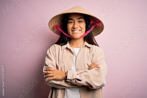 Young beautiful woman wearing traditional conical asian hat over isolated pink background happy face smiling with crossed arms looking at the camera. Positive person.